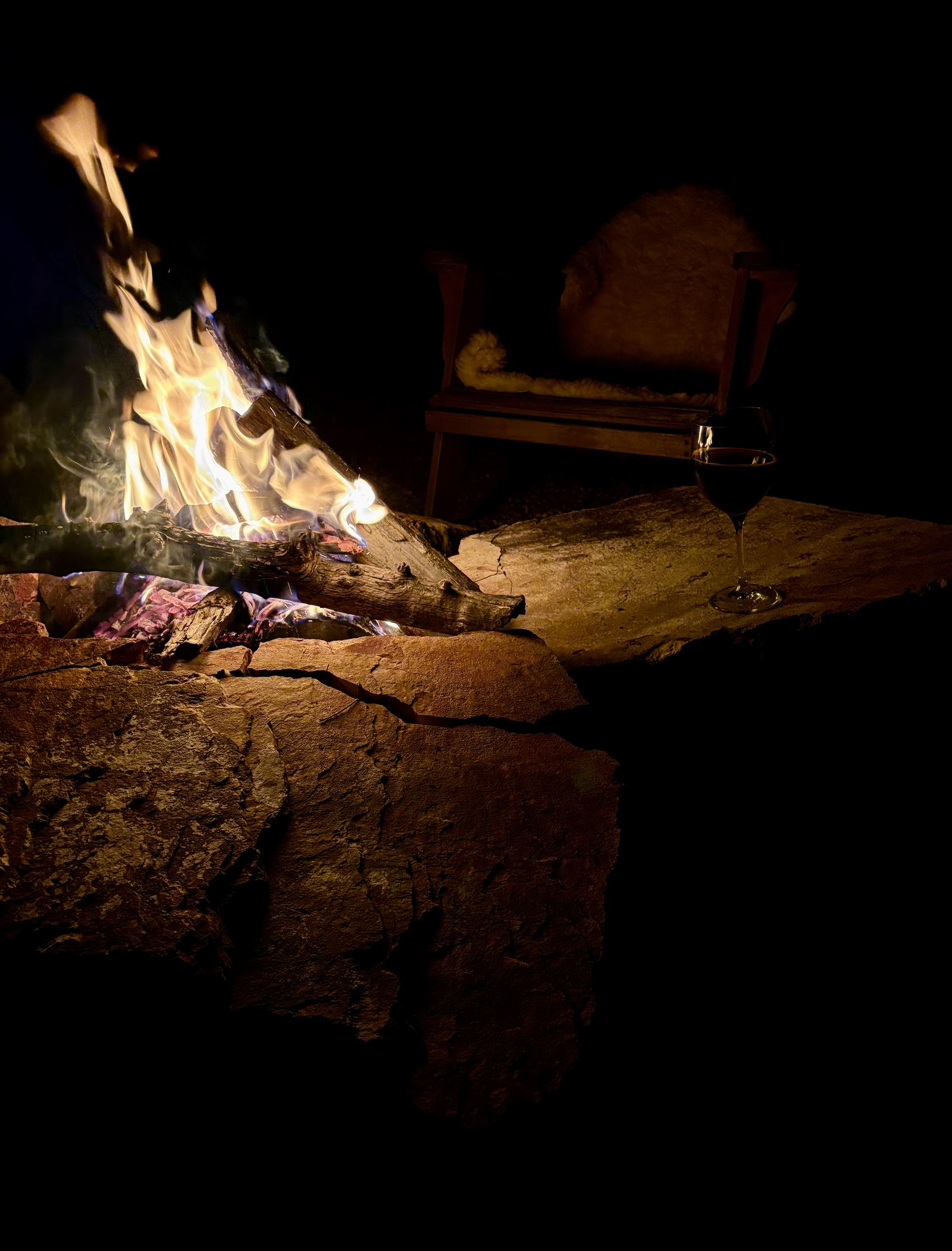 An empty wooden chair draped with a sheepskin beside a stone slab holding a glass of red wine.